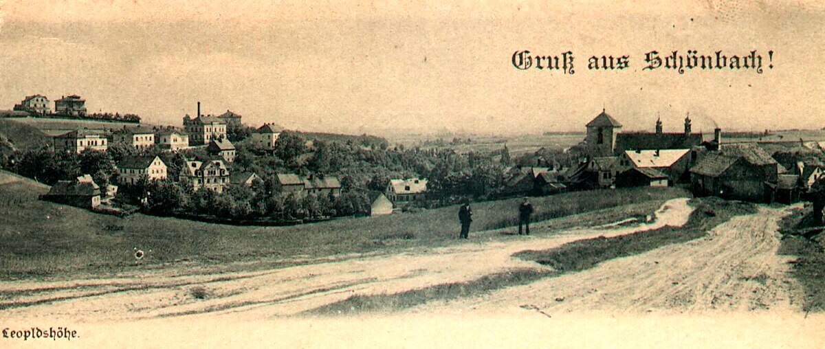 Sepia panoramic postcard titled “Gruß aus Schönbach!” and labelled Leopoldshöhe, showing a hillside village with church tower, factory chimney, clustered houses, open fields, and two small figures standing on a dirt road in the middle ground.