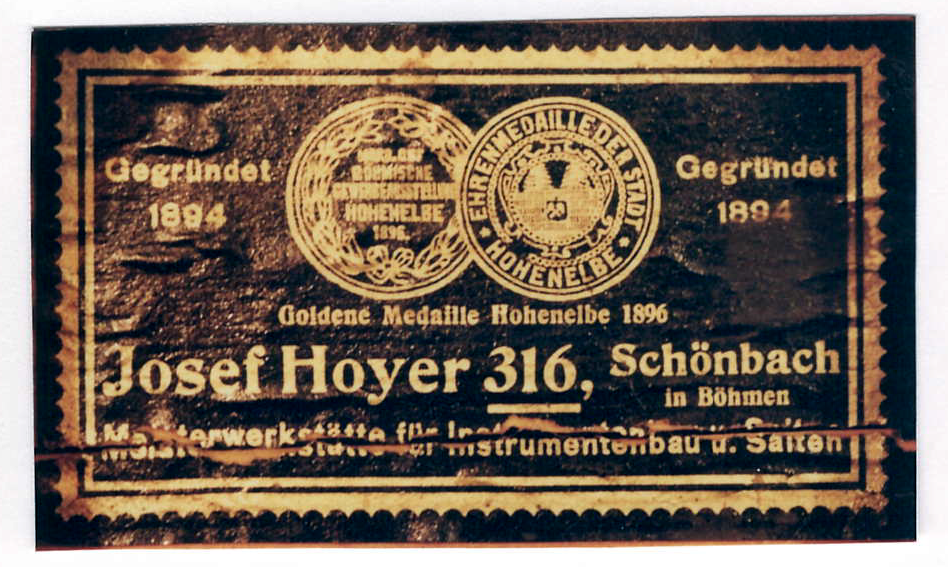 Rectangular wooden instrument label with gold lettering on dark wood: twin award seals for Hoheneibe 1896, “Gegründet 1894,” and “Josef Hoyer 316, Schönbach in Böhmen,” master workshop for instrument making and strings.