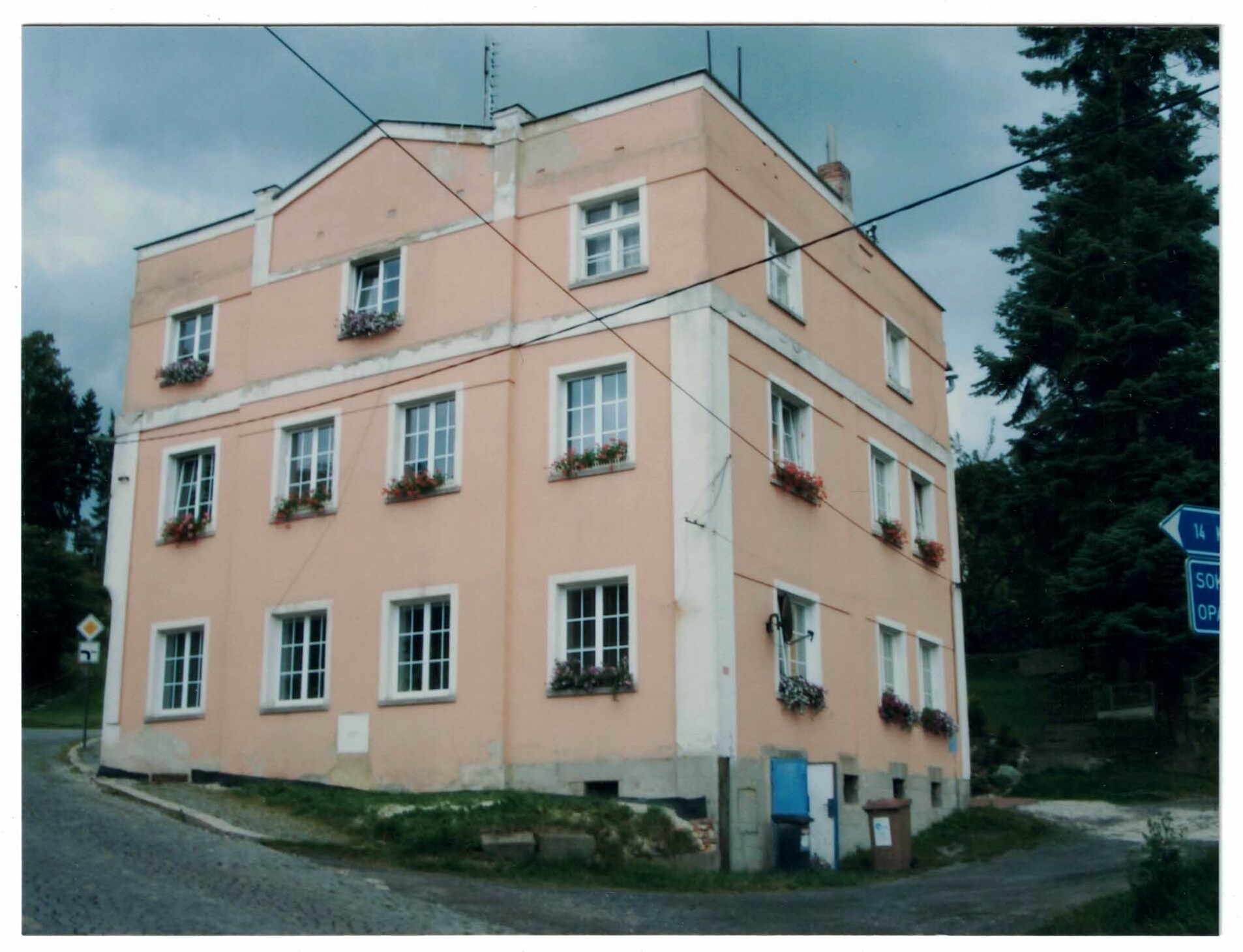Colour photograph of a three-storey peach-pink corner house with white window frames and flower boxes on a cobbled street under a cloudy sky, with a blue road sign pointing toward Sokolov.