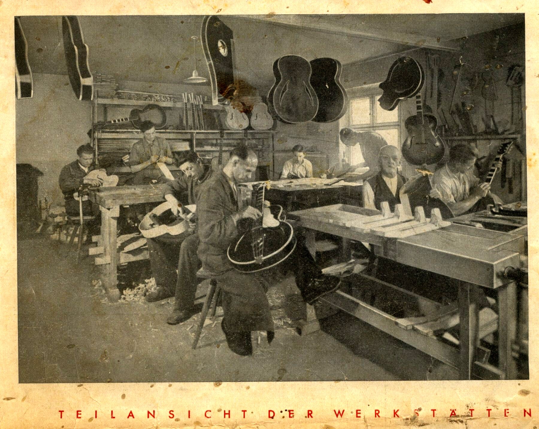 Black-and-white catalogue photograph of a busy guitar workshop with many workers at long benches, guitar bodies hanging from the ceiling and walls, and a red printed caption “TEILANSICHT DER WERKSTÄTTEN.”